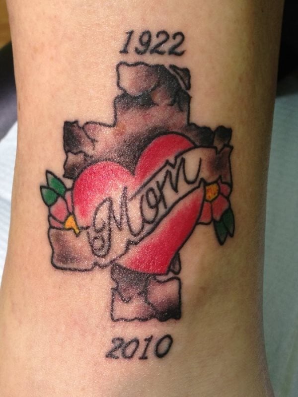 Tatuaje con la palabra mom y un corazón rodeado por unas pequeñas flores y en cuyo fondo se puede apreciar una gran cruz emulando a la piedra y la fecha 1922 tatuada en la parte de arriba