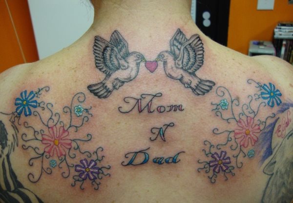 En esta ocasión a las palabras mamá y papá se le han tatuado grandes decoraciones para rodear estas palabras, como podemos ver unas peculiares flores de colores y la parte superior de la espalda se han tatuado dos pájaros iguales que portan sobre su pico un corazón