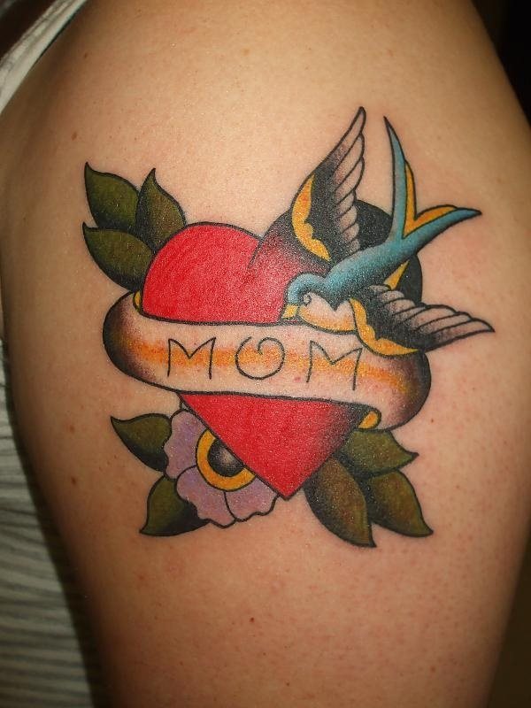 Bonito tatuaje de corazón rojo envuelto por flores y hojas con tonos suaves, custodiado el corazón por un pájaro que parece estar mirando a la palabra mamá que se ha tatuado sobre el lazo que envuelven al corazón